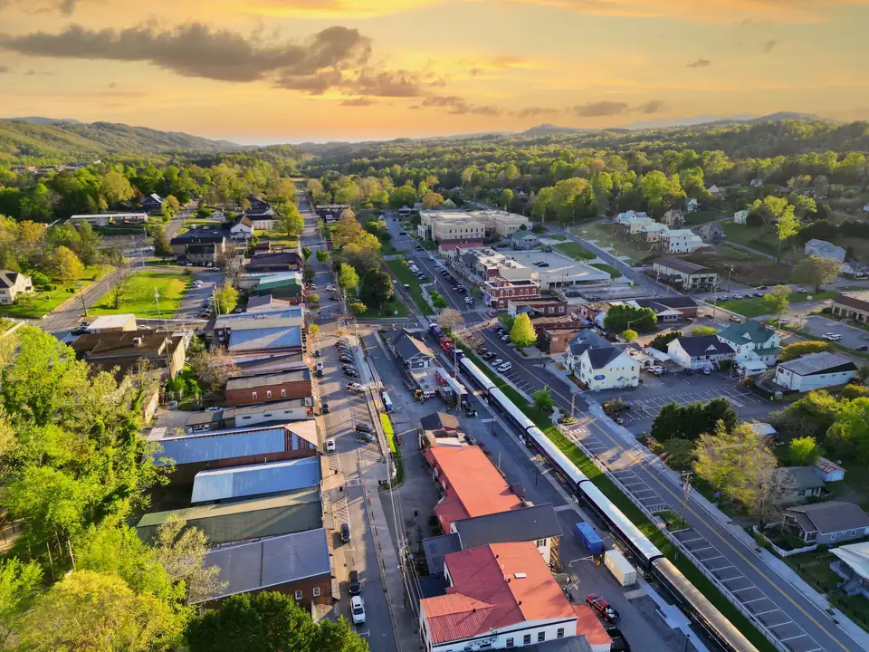 Aerial view at sunset of a small town grid with a long passenger train parked beside a depot, forested hills under an orange sky