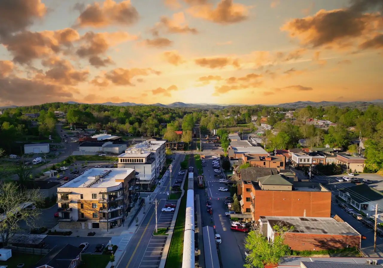 Aerial looking straight down a parked silver passenger train between two parallel downtown streets, a new four-story building at left and brick blocks at right, green hills and a golden cloudy sky