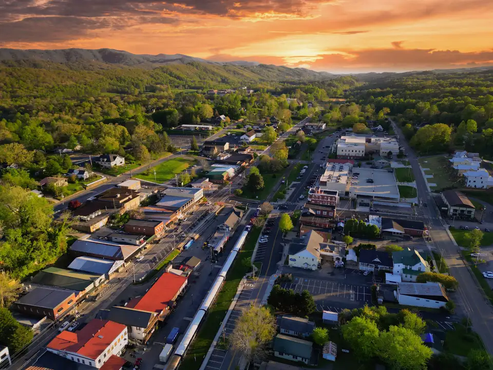 Aerial over downtown rooftops including red-roofed buildings, a long passenger train beside the tracks, streets and parking, with forested ridges and an orange sunset sky behind