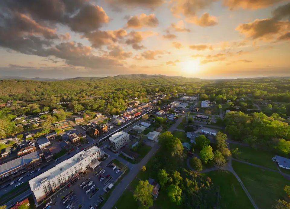 Wide aerial of the town strip and rail line running toward low mountains with the sun on the horizon, a large white multi-story building and parking lot in the foreground, green park and woods at right