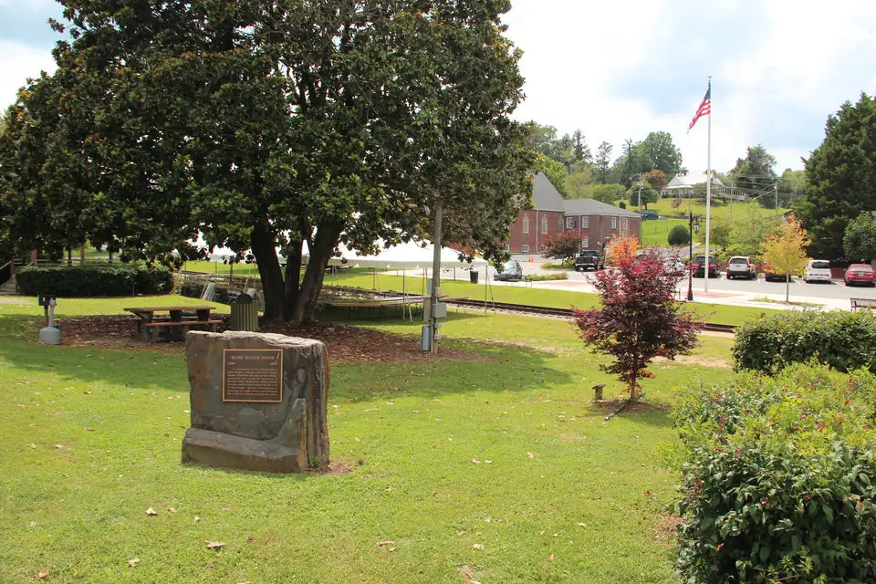 Stone marker with a bronze 'BLUE RIDGE PARK' plaque under a large magnolia, picnic table, rail track and flagpole behind