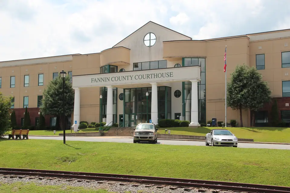 Tan multi-story building with white columns and 'FANNIN COUNTY COURTHOUSE' lettering, two cars parked, rail track in the foreground