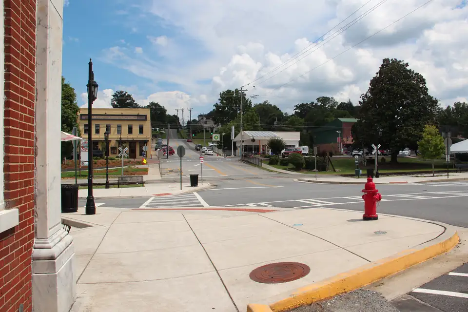 Wide sidewalk corner with a red fire hydrant and black lamp post, crosswalks, a railroad crossing sign, a two-story cream storefront building and a large magnolia under a cloudy blue sky