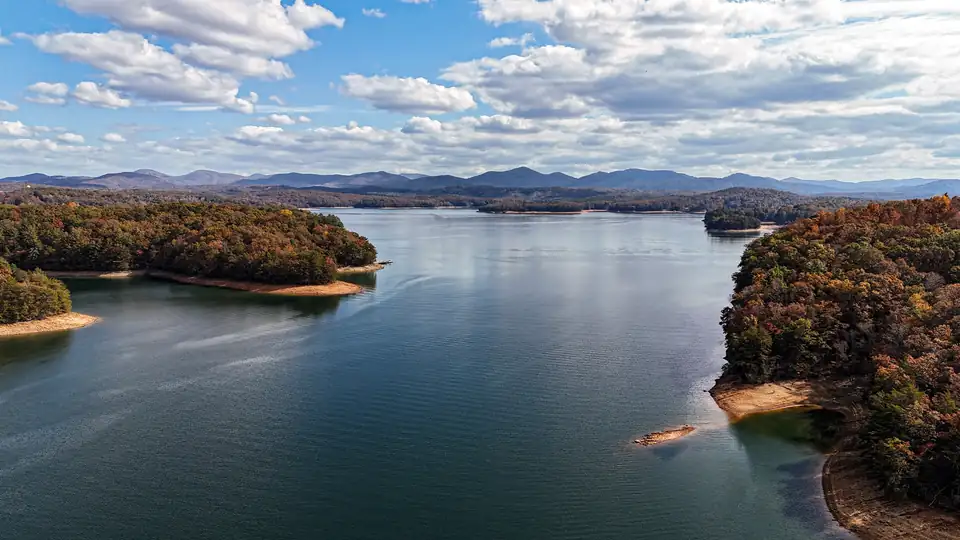 Aerial view of a wide blue lake between wooded points in orange and brown fall color, layered mountains and white clouds beyond