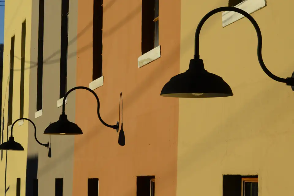 Angled close view of black gooseneck barn lamps in a row along a yellow, orange and tan painted wall with tall dark windows in warm sun