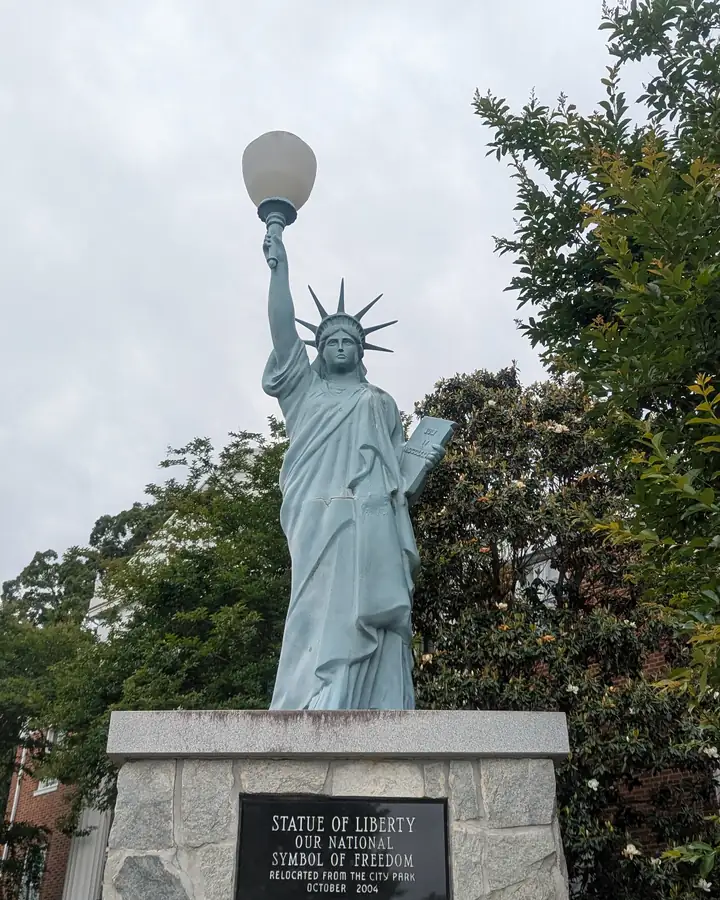 Pale green Statue of Liberty replica with a globe-lamp torch on a stone pedestal; plaque reads 'Statue of Liberty, our national symbol of freedom, relocated from the City Park, October 2004'; trees and a brick building behind under an overcast sky