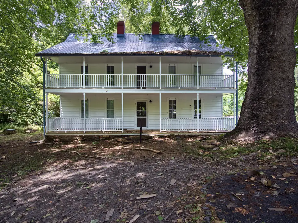Two-story white clapboard house with full-width double porches and white railings, metal roof and two red chimneys, beside a huge tree trunk, on a leaf-strewn dirt yard in green woods