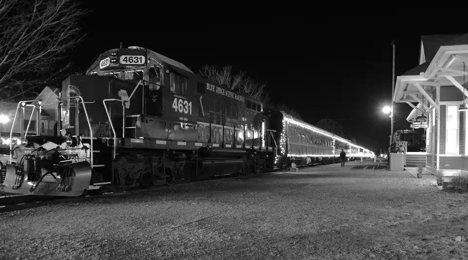 Black-and-white night view of locomotive 4631 lettered 'Blue Ridge Scenic Railway' beside a lit depot, coaches outlined with string lights