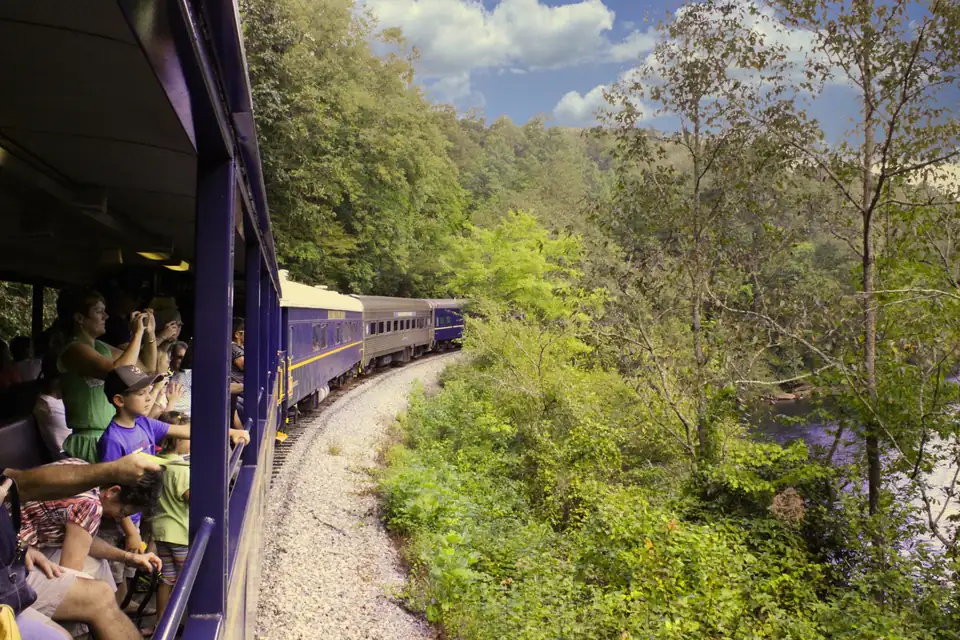 Passengers in an open-air rail car photograph the view as blue and silver coaches curve ahead through green woods beside a river