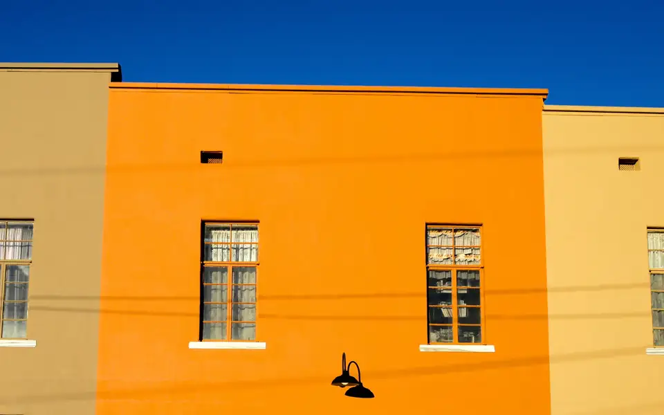 Panoramic crop of a flat-roofed building wall painted tan, bright orange and pale yellow, with six windows and black gooseneck lamps under a deep blue sky